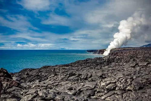 A black lava flow running to the ocean, with a steam plume rising where it meets the water