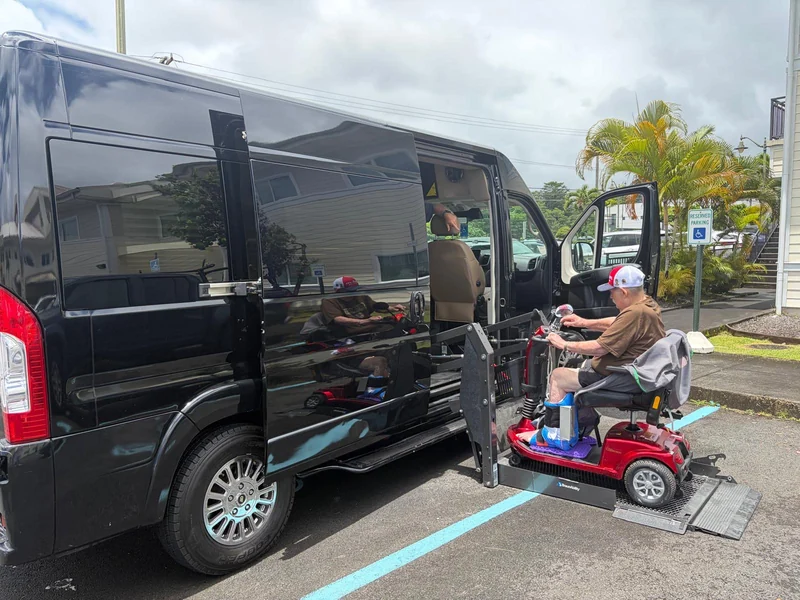 A passenger on a mobility scooter boarding the black accessible van on its hydraulic lift platform